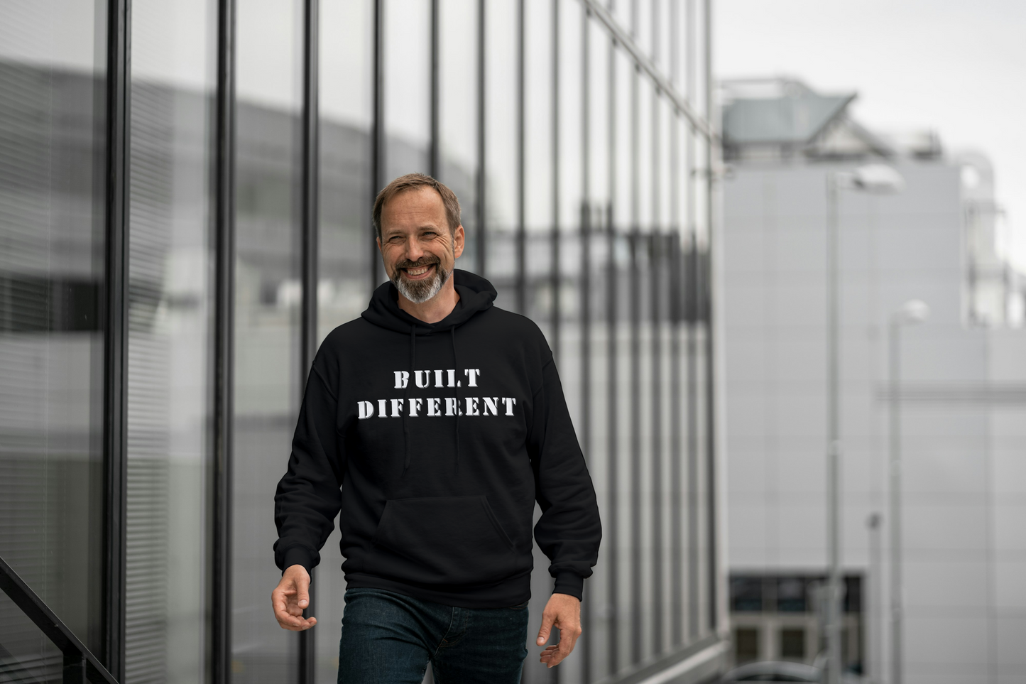 Man wearing a black hoodie with 'BUILT DIFFERENT' text, standing in front of modern glass buildings.