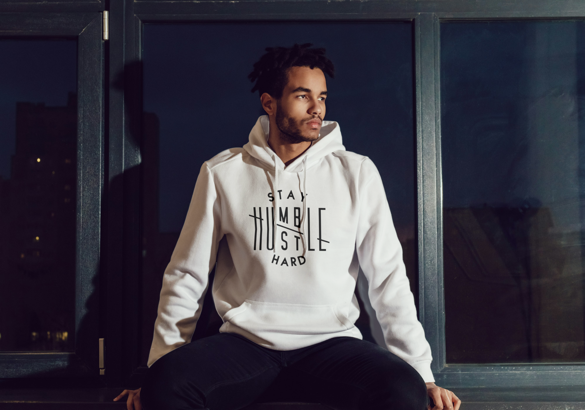Man wearing a off white hoodie with text, sitting by a window at night.